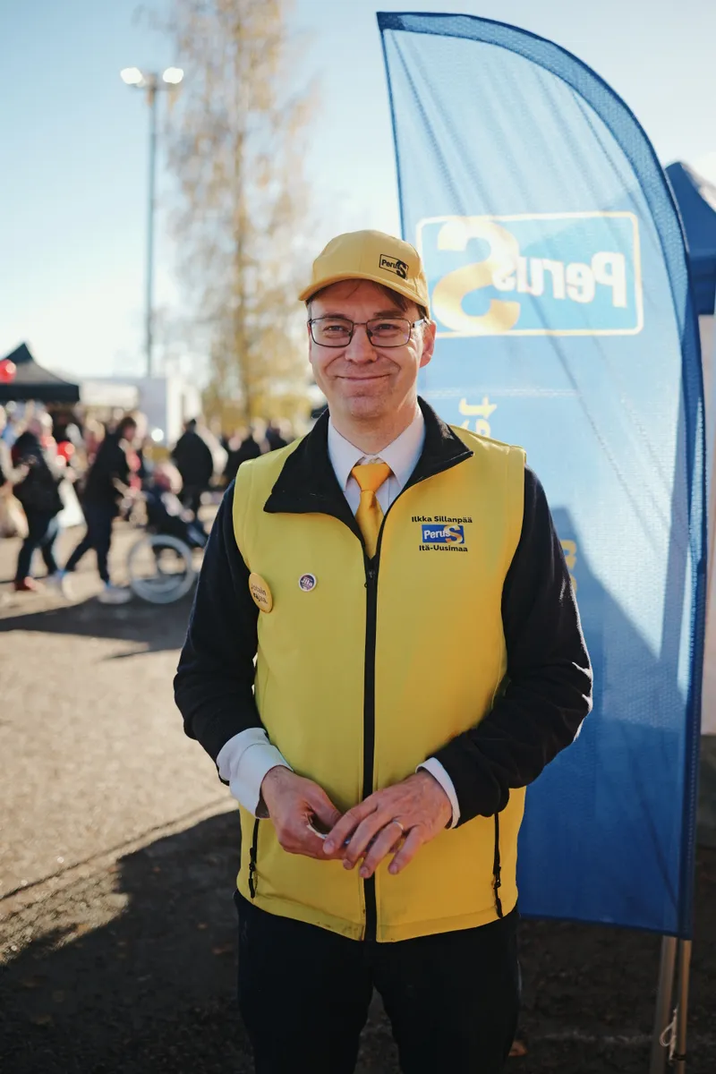Ilkka Sillanpää keltaisessa liivissä ja lippalakissa sinisen viirin edessä.